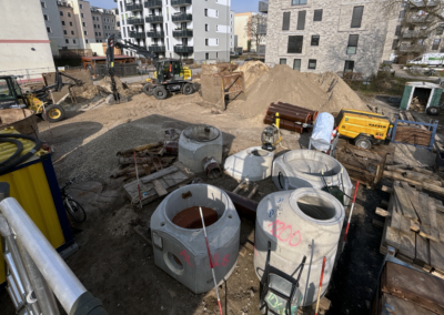 Baustelle mit mehreren großen Betonringen, Erdhaufen, Baumaschinen und mehrstöckigen Wohngebäuden im Hintergrund