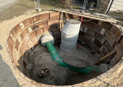 Baugrube mit runden Stahlwänden, darin ein grünes Rohr und ein zylindrischer Betonschacht mit Deckel