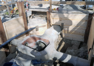 Baustelle mit einer teilweise gemauerten und betonierten Rohrleitung in einem Erdgraben, umgeben von Holzverschalungen und Baugerüsten